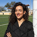 A woman with curly hair and a black jacket stands confidently by a soccer goalpost on a bright, sunny day at an outdoor field.