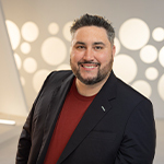 Michael La Plante smiles at the camera. He is wearing a red t-shirt with a black blazer and standing in front of a white backdrop.