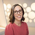 Nina Lopez-Corrado smiles at the camera. She is wearing brown glasses and a red sweater and standing in front of a white backdrop.