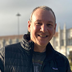 Sean Spuehler stands outdoors in front of a large building. He is smiling and wearing a black jacket and a blue t-shirt.