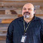 Instructor Eric Berzins, a man with a grey goatee, standing in front of a wood paneled wall wearing a blue button down dress shirt and green Full Sail lanyard with name badge.