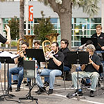 Members of the Brass Band of Central Florida perform in a parking lot, sitting down and reading from their music stands while a conductor stands at the front.