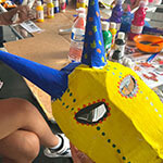 A person holds up a hand painted horned mask in front of a crowded arts and crafts table.