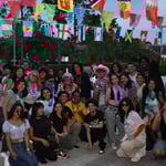 A diverse group of young adults poses outdoors under international flags, dressed festively with leis and fun accessories.