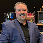 Richard Tremmel smiles at the camera with an array of music recording equipment behind him. He is wearing a black shirt and blue blazer.