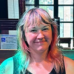 Kim Kozak sits on a piano bench in a backstage area at Garcia’s. She is smiling and wearing a black dress and tights.