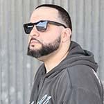 Full Sail grad George Doman wearing black sunglasses and a grey pullover hoodie with a yellow and light blue design, standing in front of a large stone wall while looking off into the distance.