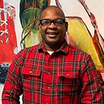 Anthony Roberts Jr. stands in front of two paintings of shoes. He is wearing black-rimmed glasses and a plaid button-down shirt.