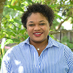 Jessica stands in front of a lush garden, surrounded by greenery. She is wearing a blue pinstripe shirt and is smiling.