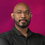 Gregg is wearing a black shirt and rectangular glasses. He is standing in front of a pink background with his arms crossed.
