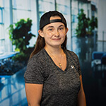 A person wearing a backward cap and a gray Under Armour shirt smiles in a bright, modern indoor space with plants in the background.