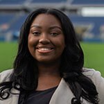 A woman with long brown hair smiling in a beige blazer while standing on a soccer pitch.