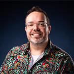 Tom Boldman smiles at the camera in front of a dark background. He is wearing a colorful paisley  shirt and glasses.