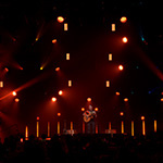 Trivium lead-singer Matt Heafy on a dimly-lit stage playing an acoustic guitar in front of a large crowd.