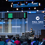 A person is seated at a desk on a stage in front of a crowd of students while a large LED screen behind displays a mirror of the audio program running on their computer.