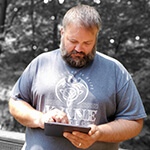 Grad Ryan Backa in a blue shirt with a white dragon design, holding an electronic tablet in front of a wooded area in greyscale.