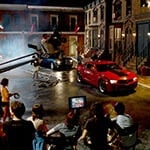 Full Sail University's Backlot in the evening filled with a full film production crew shooting a scene featuring two muscle cars, one red and one black.