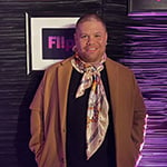 Javi Simons stands in front of a purple wall. He wears a black t-shirt, jeans, a brown overcoat, and a colorful scarf. He is smiling.