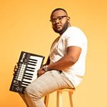 Chavez Parker sits on a stool and holds a keyboard. He wears glasses, a t-shirt, brown pants, and sneakers.