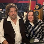 Alan Parsons sits with Steffie Tjandra in front of a mixing board in a recording studio. They are smiling at the camera.