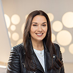 A headshot of a woman with straight brown hair and hoop earrings smiling while wearing a leather motorcycle jacket against a white background.
