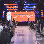 A bustling career fair with several booths set up. In the center, a giant LED screen welcomes guests to the Career Fair.