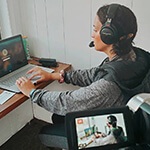 Grad Sydney Phillips, a woman with dark brown hair wearing a grey hooded sweatshirt and black over-ear headphones, is seated at a desk on her laptop in a press box overlooking athletes in uniform on a football field.