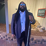 Andre Woods stands outdoors in front of a restaurant. He is wearing a blue suit and sunglasses. He is holding a mobility cane.