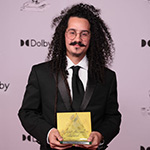 Guillermo Moya smiles at the camera in front of a white background. He is wearing a black suit, black tie, and glasses.