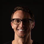 Drew Becker smiles at the camera in front of a dark background. He is wearing black glasses and a black shirt.