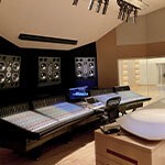 A conference table faces a wall with mixing boards and speakers; a recording area with a grand piano is in the background.