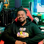 Grad Shawn Deshield smiling while seated at a desk in a home studio featuring a keyboard, audio equipment, and a large screen displaying audio software while wearing a black hooded sweatshirt with a colorful design.