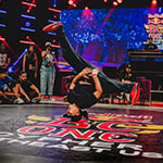 A dancer is doing a hands-free headstand with their legs spread while their competitor, the audience, and the judges watch.