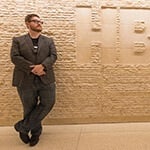 Wearing a suit and glasses, Stephen Beres leans against a beige, textured stone wall with the HBO logo carved into it.