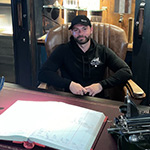 Rob Prager sits in a leather chair behind a large desk. He is smiling and wearing a black baseball cap and a black hoodie.