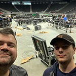 Two smiling men with beards stand in a backstage area. There are wires on the floor behind them, as well as an open trunk filled with production equipment.