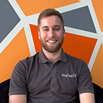 A man with light hair and a beard wearing a grey Holovis polo while seated and smiling in front of an orange, grey, and white geometric background.