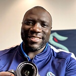 Grad Lamont Buford sports a blue jacket with a Seattle Kraken logo while smiling and holding a camera.