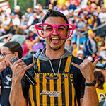 A man standing and looking excited while holding a microphone in oversized pink glasses and yellow and black striped overalls in the foreground of a large crowd at a stadium.