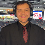 A smiling sports broadcaster in a suit and headset stands in an arena booth above an ice hockey rink with "Game Day" on screens behind.