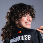 A smiling woman with curly hair holds up a jersey that reads “miamouse” on the back, showing it proudly to the camera.