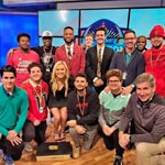 A group shot of several Full Sail students and instructors in front of a large screen on set at the Dan Patrick School of Sportscasting.