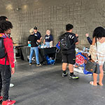 Several students gather on a patio with various tables set up. Club representatives stand at the tables talking with students.