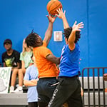 Two basketball players jump up simultaneously to reach the basketball on a court. They are both wearing athletic clothing.