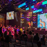 Several students raise their hands attentively at the Summer Sizzle as two staff members call on participants. The large room is colorfully lit up and looks very lively.