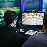 Two people have their backs to the camera while watching ‘NBA 2K’ gameplay on a small monitor on a table in front of them.