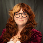 Susan Kost smiles at the camera. She has long red hair and is wearing a white blouse, a red velvet jacket, and black glasses.