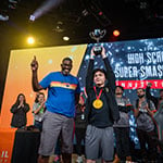Two people are standing side by side on a stage while one holds a large silver trophy over their head.