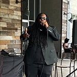 Guest speaker Souljah Bless, dressed in an all black outfit, speaks into a microphone on a patio stage. Behind him, a DJ stands at a booth that reads “Student Community Association.