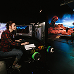 A woman is sitting at a desk in front of several monitors. She is in a virtual production studio that has been designed to look similar to Mars.